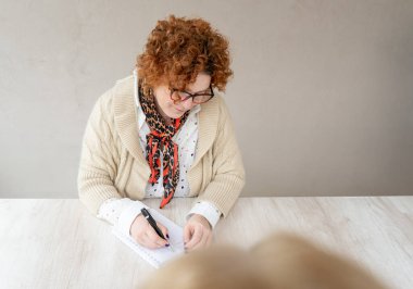 Düşünceli, orta yaşlı, kıvırcık saçlı, gözlüklü, eşarp takan ve rahat bir süveter giyen bir kadın, ahşap bir masada otururken sarmal bir not defterine notlar yazarken yakalandı.