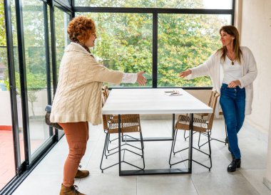 İki orta yaşlı kadın, büyük pencereli ve minimalist dekorlu şık, aydınlık bir odada birbirlerini hoş bir jestle selamlıyorlar.