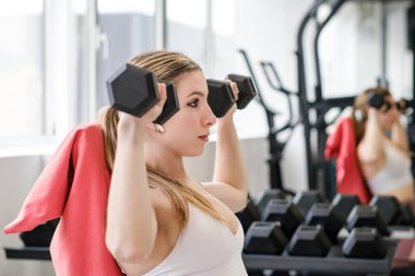 Odaklanmış bir spor salonunda, bir kadın omuzlarını dambıllarla bastırarak vücudunun üst kısmını hedef alır.