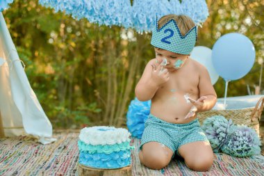 In a charming outdoor setting, a young toddler partakes in a cake smash, playfully eating and exploring a blue and white iced cake during his second birthday celebration clipart