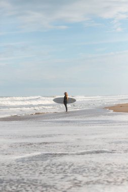 Eğitim sırasında ıslak kıyıda dalgalı deniz kenarında duran siyah dalış kıyafetli uzak sportif kadın sörfçünün tüm vücudu.