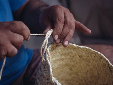 Yakından çekilen görüntü, palmito iplikçikleriyle bir sepet ören yetenekli bir zanaatkârın detaylı çalışmasını içeriyor.
