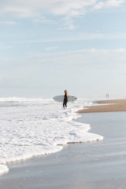 Eğitim sırasında ıslak kıyıda dalgalı deniz kenarında duran siyah dalış kıyafetli uzak sportif kadın sörfçünün tüm vücudu.
