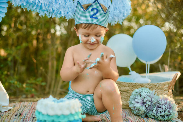 Радостный малыш в сине-белой короне наслаждается разбитым тортом во время празднования своего второго дня рождения на открытом воздухе, окруженный воздушными шарами и цветами