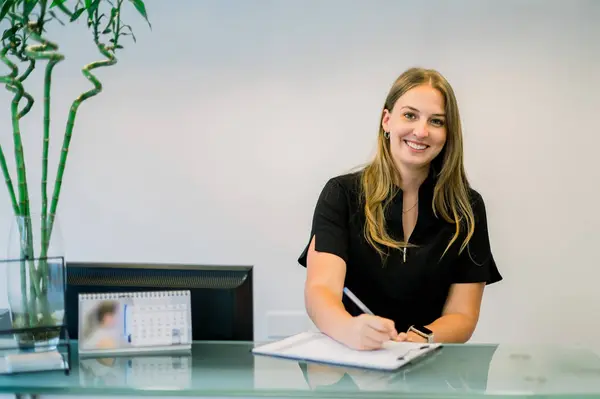 Siyah üniforma giymiş gülümseyen bir resepsiyonist şık cam bir masanın arkasında oturup estetik bir klinikte hastaları selamlıyor elinde hasta formlarına yardımcı olmak için bir not tahtası ve kalem tutuyor.