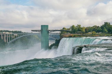 Striking panoramic shot of Niagara Falls, showcasing the powerful cascades with the iconic Rainbow Bridge connecting the USA and Canada, set against a cloudy sky clipart