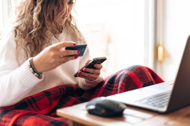 Genç kıvırcık saçlı, beyaz kazaklı bir kadın bilgisayarlı masada otururken internet üzerinden akıllı telefon ve kredi kartı kullanıyor.