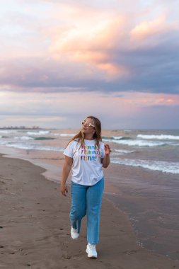 Moda güneş gözlüklü ve şık kıyafetli genç bir kadın yaz akşamları deniz kenarında yürüyor.