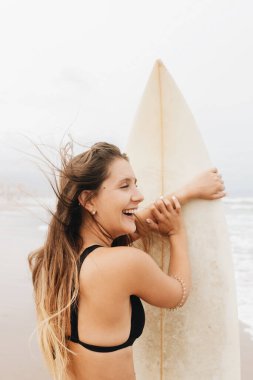 Side view of positive young female athlete in swimsuit with flying hair and surfboard looking away on ocean beach clipart