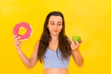 Bir kadın bir elinde pembe jöleli bir çörek, diğer elinde yeşil bir elma tutuyor ve canlı sarı bir arka plana karşı seçim yapıyor..