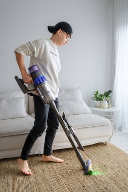 Genç bir birey rahat bir oturma odasında kablosuz elektrik süpürgesiyle halıyı süpürmeye odaklanır.