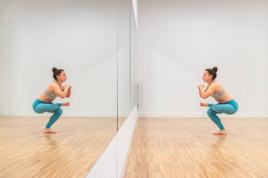 Deniz mavisi yoga pantolonlu kadının yan görüntüsü, sakin bir yoga stüdyosundaki aynanın önünde Garland Poose (Malasana) sergilemektedir..