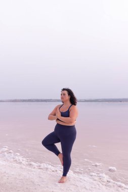 Pembe gölün kıyısında Vrksasana 'da dikilip yoga yapan hareketli kıyafetli sakin bir kadın.