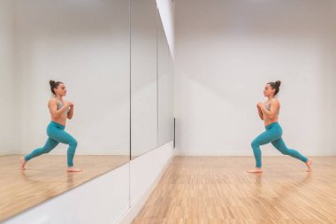 Side view of focused woman performs the Anjaneyasana yoga pose in front of a mirror in a minimalist studio. clipart