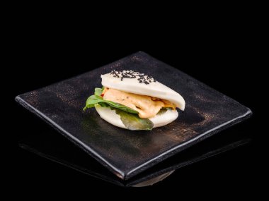 Bir gurme bao sandviçi, koyu bir tabakta zarifçe sunulur. Sandviç taze yapraklı yeşillikler ve susam tohumlarıyla kaplı kremalı bir sos içerir.