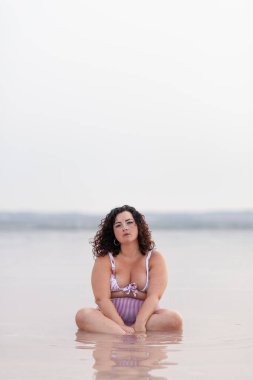 Ciddi kıvrımlı, bikinili bir kadın yazın pembe gölette oturuyor ve kameraya bakıyor.