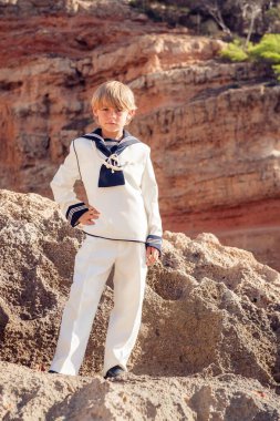 Geleneksel denizci kıyafeti giymiş genç bir çocuk kayalık arazide duruyor ve doğal bir zemine karşı ilk komünyonunu kutluyor..