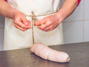 Kasap elleri, taze bir Sobrasada sosisini iple bağlamak, temiz bir endüstriyel mutfak zeminine karşı geleneksel sosis yapma tekniklerini detaylandırmak.