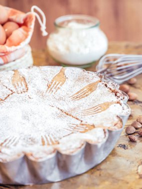 Taze pişmiş bir turta, dekoratif şeker katmanıyla kaplı metal bir kabın içinde oturur. Arka planda bir çırpı, bir kase un ve yumurta bu kırsal pişirme sahnesini tamamlıyor.