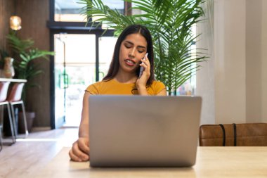Kafedeki ahşap masada oturan ve netbook 'ta yazarken telefonla konuşan ciddi bir serbest çalışan kadın.