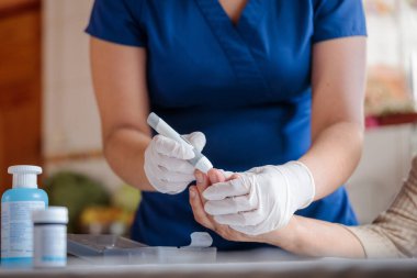 Eldiven takan bir hemşire, yaşlı bir hastanın kan şekerini ölçmek için bir glukometre hazırlar ve evdeki glisemiyi kontrol etmeye çalışır.