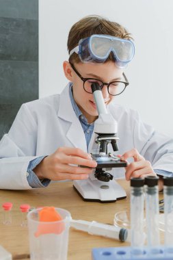 Çocuk üniformalı, test tüpünü mikroskopla inceliyor. Masada oturuyor ve okuldaki laboratuvarda araştırma için hazırlanıyor.