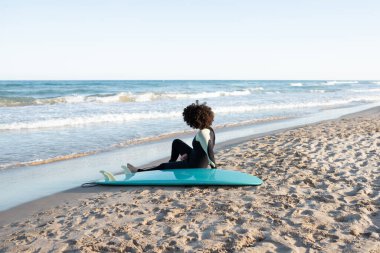 Arkadan bakıldığında çıplak ayaklı, dalgıç kıyafetli, kumlu sahilde el sallayan kadın sörfçü.