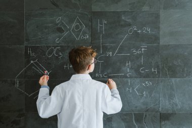 Gözlüklü ve beyaz üniformalı bir öğrencinin sınıftaki karatahtaya kimyasal formüller yazışının arkası.
