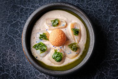 Haşlanmış marul, kızarmış peynir topu ve mavi damlalarla süslenmiş deniz tarağı ile süslenmiş iştah açıcı krem çorbanın en üst görüntüsü.