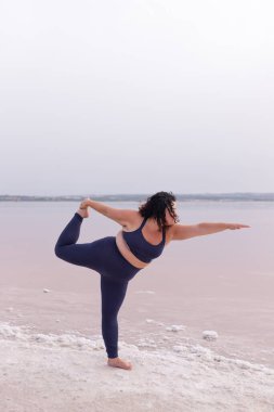 Natarajasana 'da, pembe göletin kıyısında spor kıyafetleriyle dengede duran ve yaz aylarında yoga yapan kıvrımlı bir kadın.