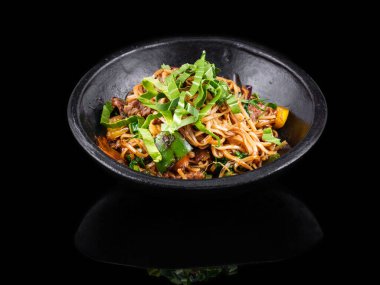 Lezzetli bir Asya usulü kızarmış et ve eriştenin üzerine karışık sebze dilimleri, taze yeşil otlarla süslenmiş, koyu siyah bir fonda sunulan, nefis bir servis.