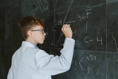 Gözlüklü ve beyaz üniformalı bir öğrencinin sınıftaki tahtaya kimyasal formüller yazışının yan görüntüsü.