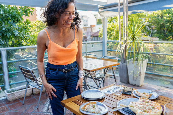 A joyful mid-adult woman laughs while standing at a dining table outdoor, featuring pizzas and table settings, depicting a casual get-together with friends on a sunny balcony