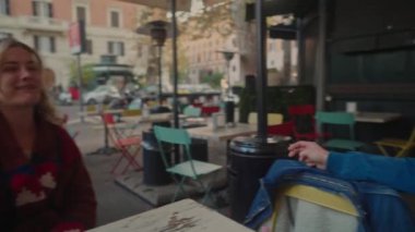 Mavi bereli ve ceketli neşeli bir kadın ve genç bir adam kalabalık bir şehir ortamında açık bir kafede otururken gülüyorlar, şehir yaşam tarzını gösteriyorlar.