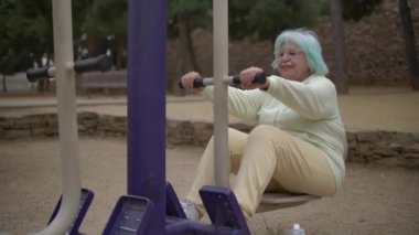 Açık yeşil ceket ve sarı pantolon giyen, mavi saç egzersizi yapan, neşeli bir ifadeyle kameradan uzağa bakan neşeli, yaşlı bir kadın.