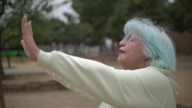 Turkuaz mavi saçlı son sınıf öğrencisi bir kadın kollarını öne uzatıyor. Açık havada, ağaçlarla kaplı, huzurlu bir parkta, gözlüklü ve açık renk ceketli tai chi çalışırken.