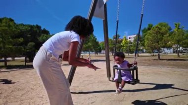 Afrikalı bir Amerikalı anne, genç kızını sevgi dolu bir şekilde güneşli bir parkta salıncakta sallıyor ve neşeli bir aile anını vurguluyor.