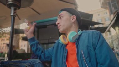 Mavi ceketli ve bereli genç bir adam, bir şehir kafesinde, kulağında kulaklıklarla, düşünceli bir şekilde oturuyor.