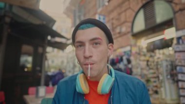 Canlı bir tarzı olan genç bir adam, mavi kulaklık takıyor, mavi saçlı ve siyah şapkalı, sigara içiyor ve gülümsüyor, kentin gençlik kültürünü somutlaştırıyor.