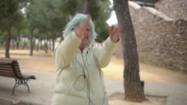 Sarı kapüşonlu, mavi saçlı yaşlı bir kadın, parkta egzersiz yaparken kollarını kaldırıyor, ileri yaşta canlılık ve sağlığını gösteriyor.