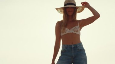 Hasır şapkalı bir kadın plajda huzur içinde duruyor. Gün batımının altın parıltısında güneşleniyor. Işıklar sakin sahneyi aydınlatıyor..