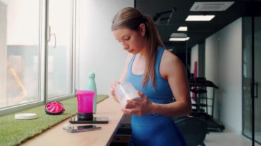 Spor salonunda odaklanmış bir bayan sporcu protein içeceği hazırlıyor, spor malzemelerinin yanında takviye tozu ölçüyor.