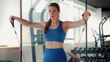 Mavi spor sutyen ve tayt giyen odaklanmış genç bir kadın direnç bantlarıyla iyi donanımlı bir spor salonunda egzersiz yapıyor. Kararlılık ve güç gösteriyor.