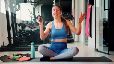 Mavi spor takım elbiseli genç bir kadın spor salonundaki yoga minderinde oturuyor, telefonuyla şarkı söylüyor ve dans ediyor, spor aletleri ve kişisel eşyalarıyla çevrili.