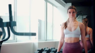 Modern bir spor salonundaki odaklanmış genç bir kadın dambıllarını kaldırarak spor kıyafeti ve kulaklık takıyor.