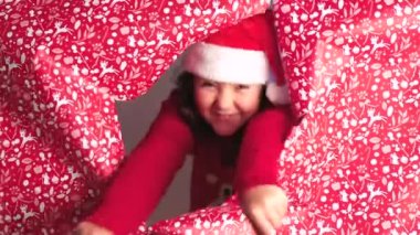 Noel Baba şapkası takan neşeli genç bir kız ve parlak kırmızı bir kazak Noel ruhunu kutluyor, kırmızı-beyaz desenli Noel ambalajı kağıdının yanında şakacı bir şekilde dikizliyor.