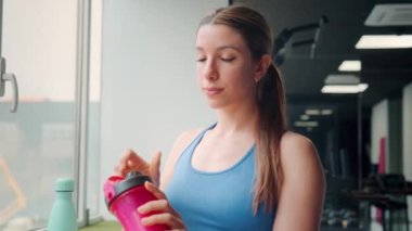 Formda bir kadın egzersiz sırasında susuzluğunu gösteren iyi donanımlı bir spor salonundaki pembe bir içecekten içmek için antrenmanını durdurur.