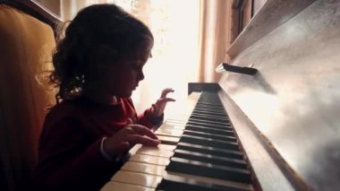 Genç bir kız, kıvırcık saçlı, piyanoyla meşgul, parmakları tuşların üzerine yayılmış, sıcak ışıklı bir odada, katıksız perdeli bir pencerenin yanında.