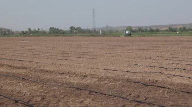 Bir kırsal çiftlik sahnesi açık bir gökyüzü altında soğan ekimine hazır bir tarla gösteriyor, ekin ekimi ve damlatma sistemleri var