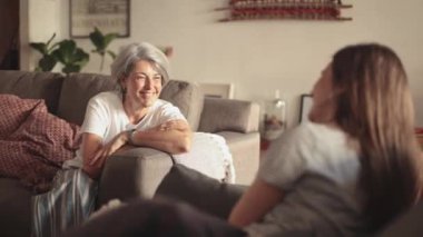 Orta yaşlı, gümüş saçlı bir anne, genç kızıyla oturma odasında rahat bir sohbet sırasında sıcak bir şekilde gülümsüyor.